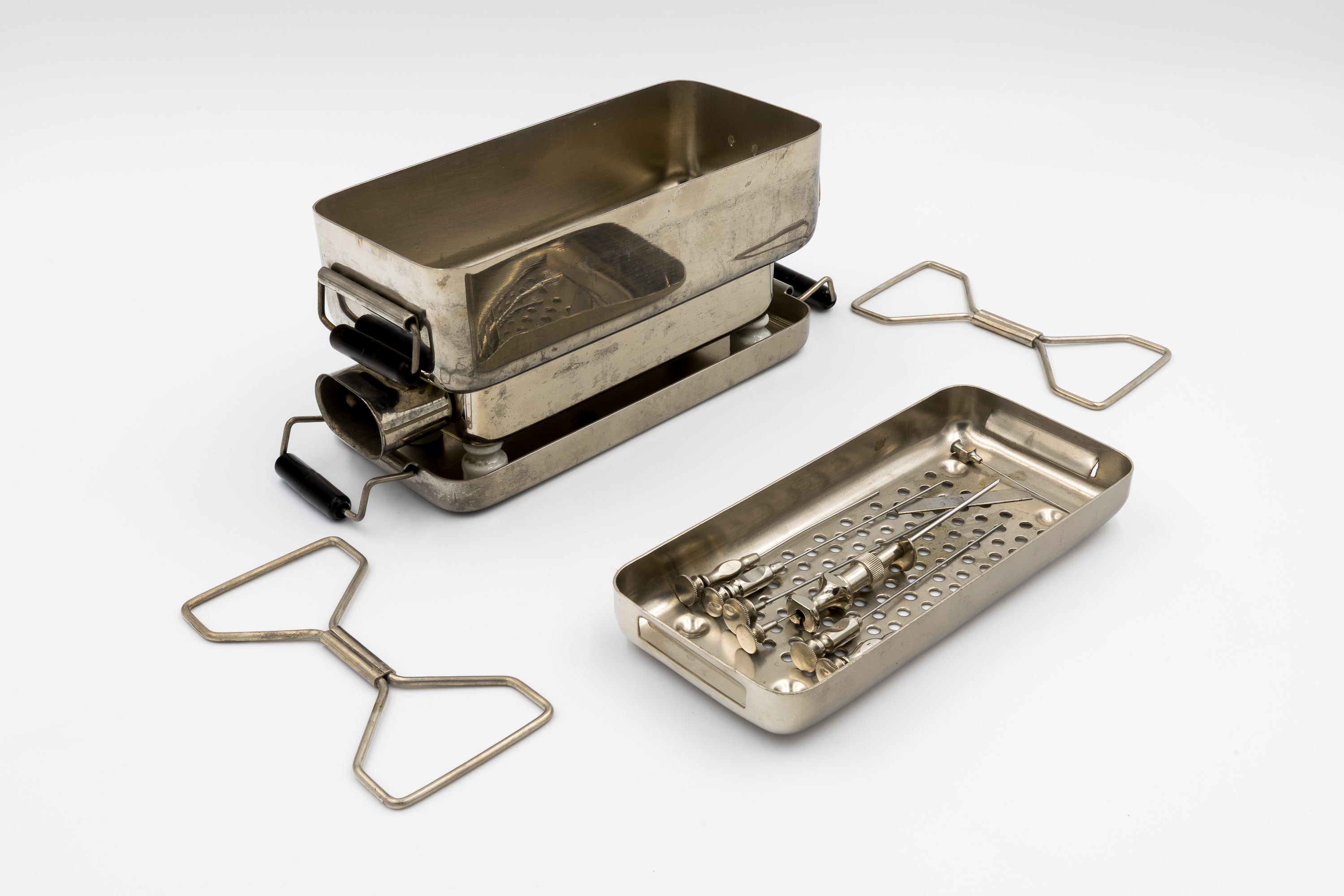 Metalowy inhalator elektryczny przeznaczony do użytku domowego. Urządzenie ma cylindryczny korpus wykonany z niklowanej stali, z czarnym uchwytem i metalową pokrywą. Na jego szczycie znajduje się metalowa rurka z żółtym przewodem i szklaną rurką do odprowadzania pary wodnej lub aerozolu leku. Obok aparatu stoi szklany pojemnik. Całość ustawiona jest na białym tle.